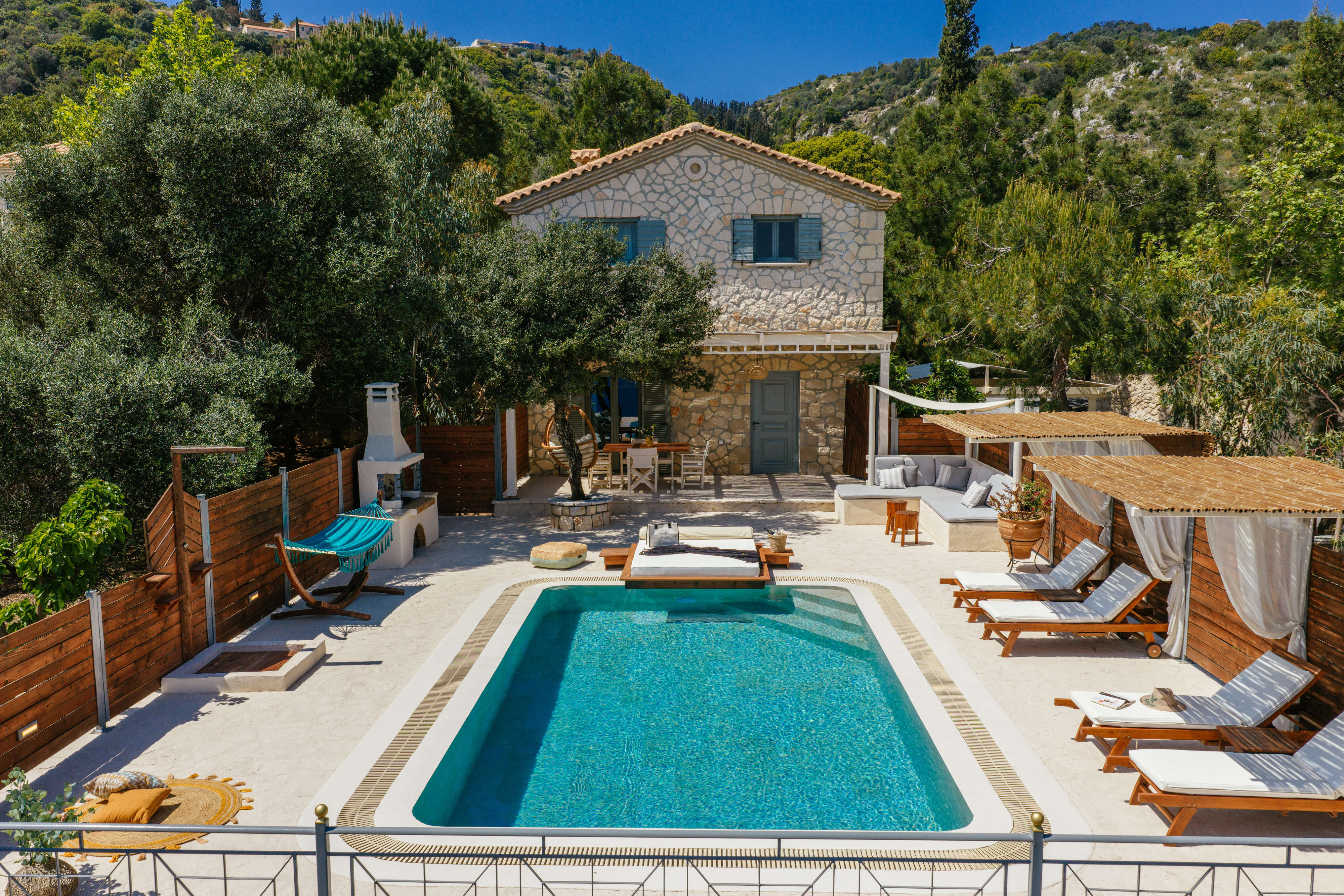 Elevated view of the stone villa nestled among olive and pine trees, with the pool, hammock, and shaded terrace seating visible below.