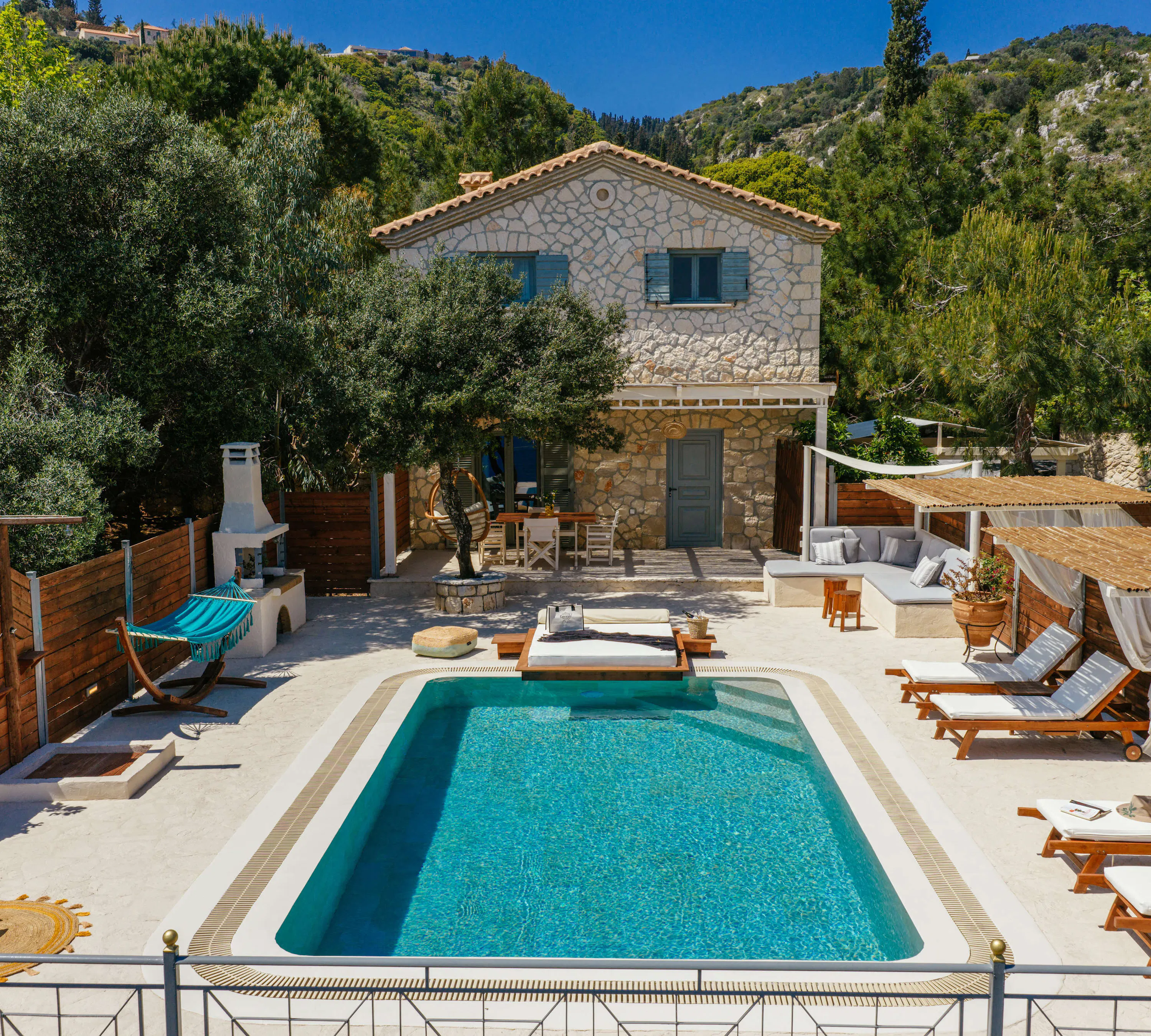 Elevated view of the stone villa nestled among olive and pine trees, with the pool, hammock, and shaded terrace seating visible below.