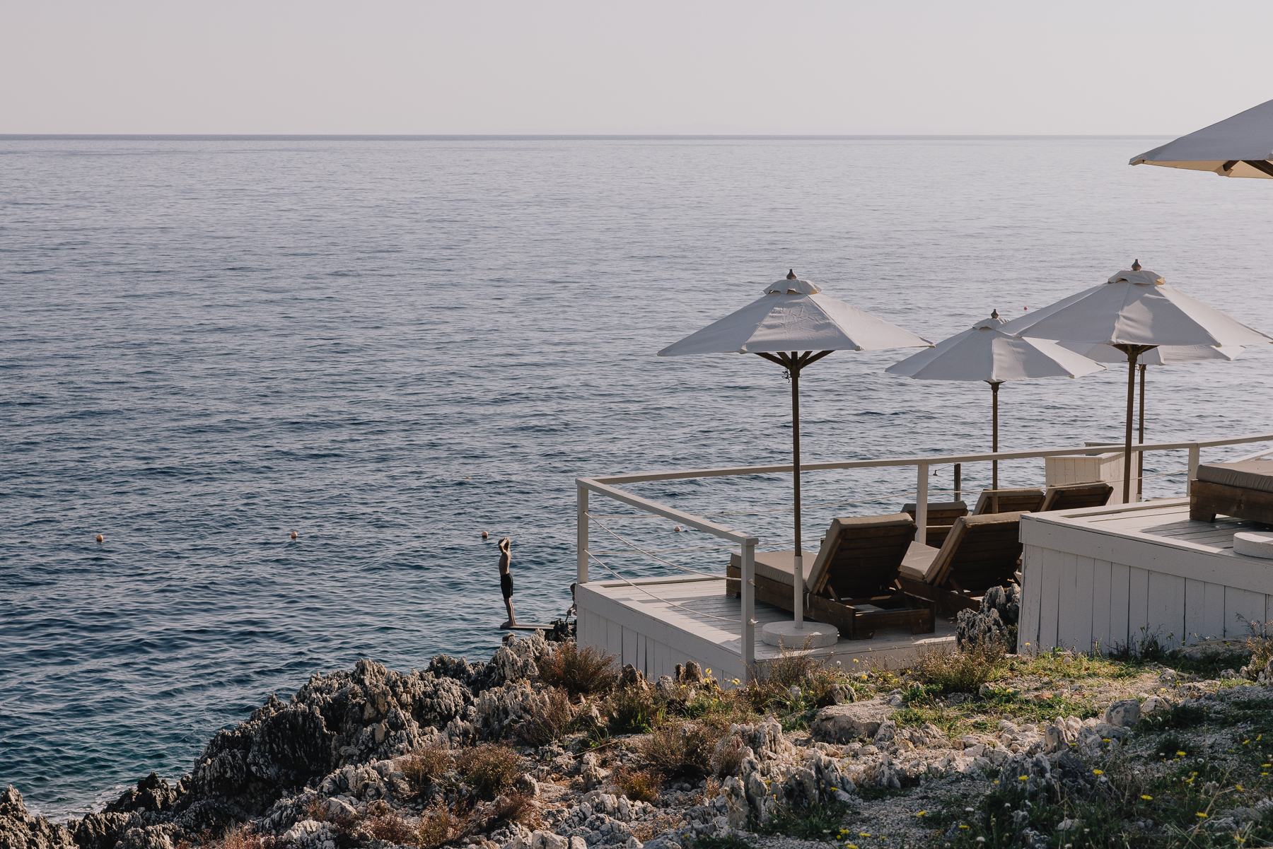 A quiet cliffside deck area with three cream umbrellas shading wooden loungers overlooks the sea, a diving platform with a lone swimmer standing at its edge visible on the rocky outcrop below against a hazy horizon.