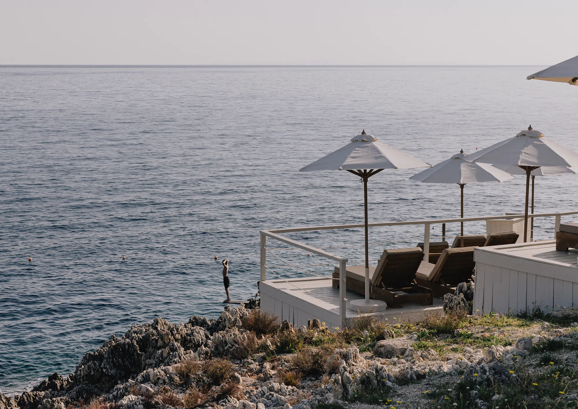 A quiet cliffside deck area with three cream umbrellas shading wooden loungers overlooks the sea, a diving platform with a lone swimmer standing at its edge visible on the rocky outcrop below against a hazy horizon.