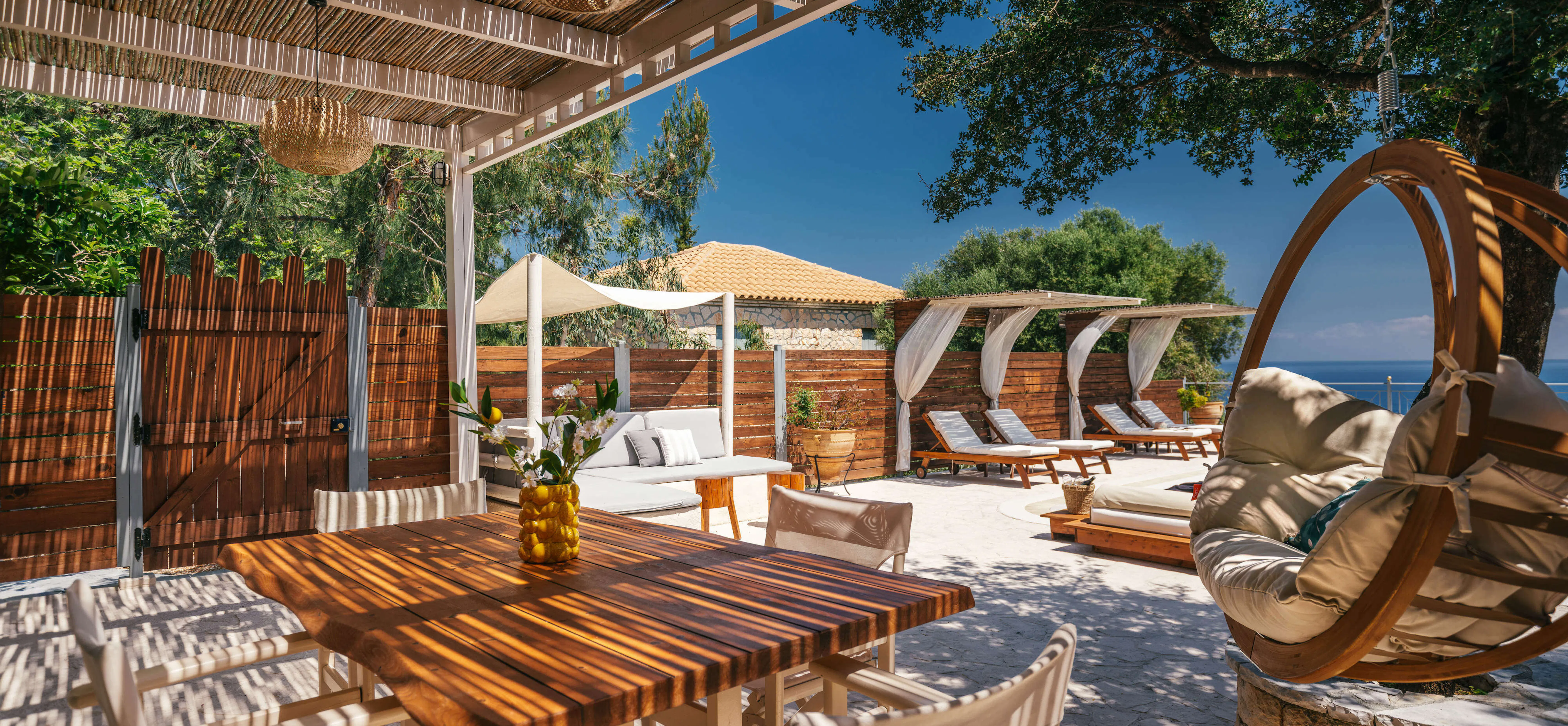 Shaded pergola dining and lounge area overlooking the pool, with a wooden table, striped director's chairs, and a hanging wicker egg chair beside a stone fire pit.