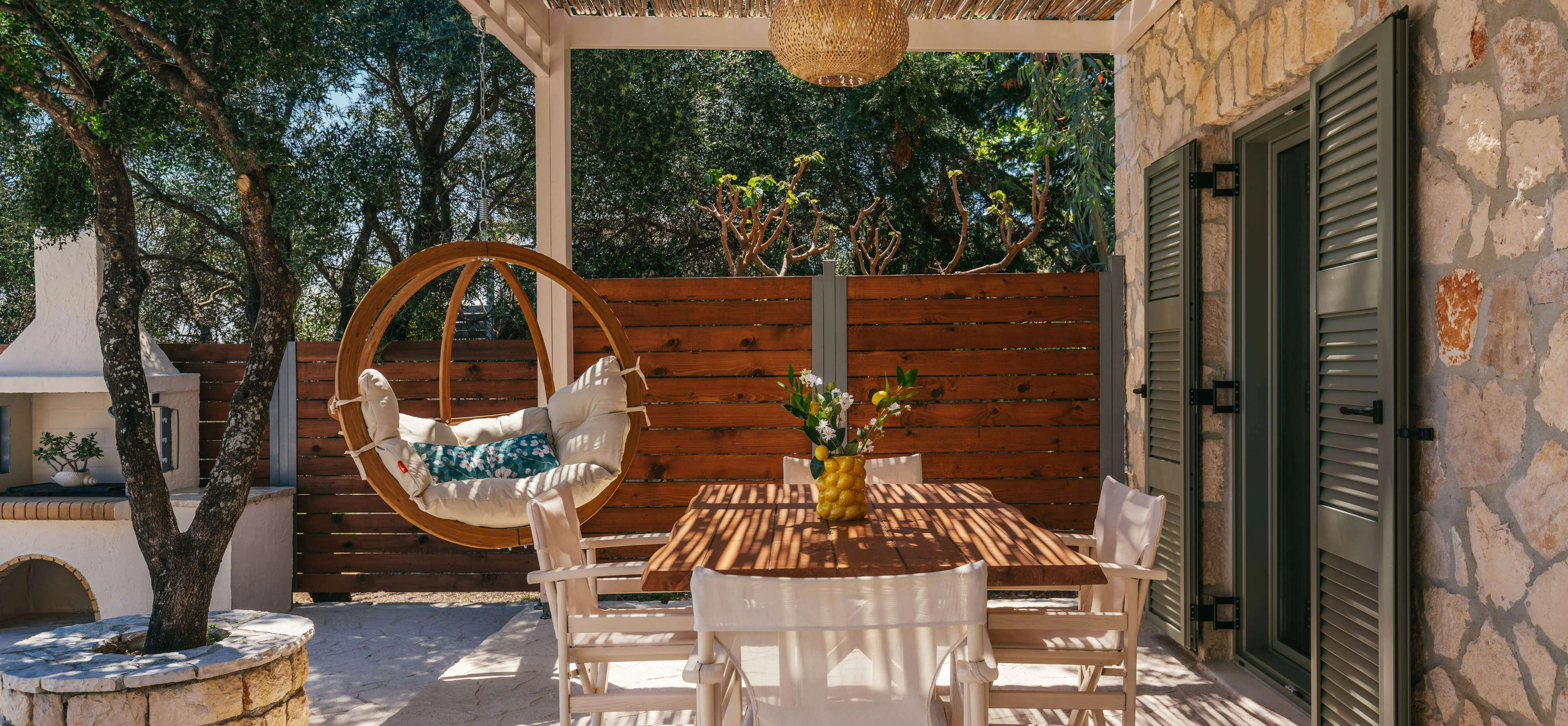 Shaded outdoor dining terrace with a rustic wooden table, white director's chairs, a hanging egg chair, and a stone-built barbecue against a backdrop of olive trees.