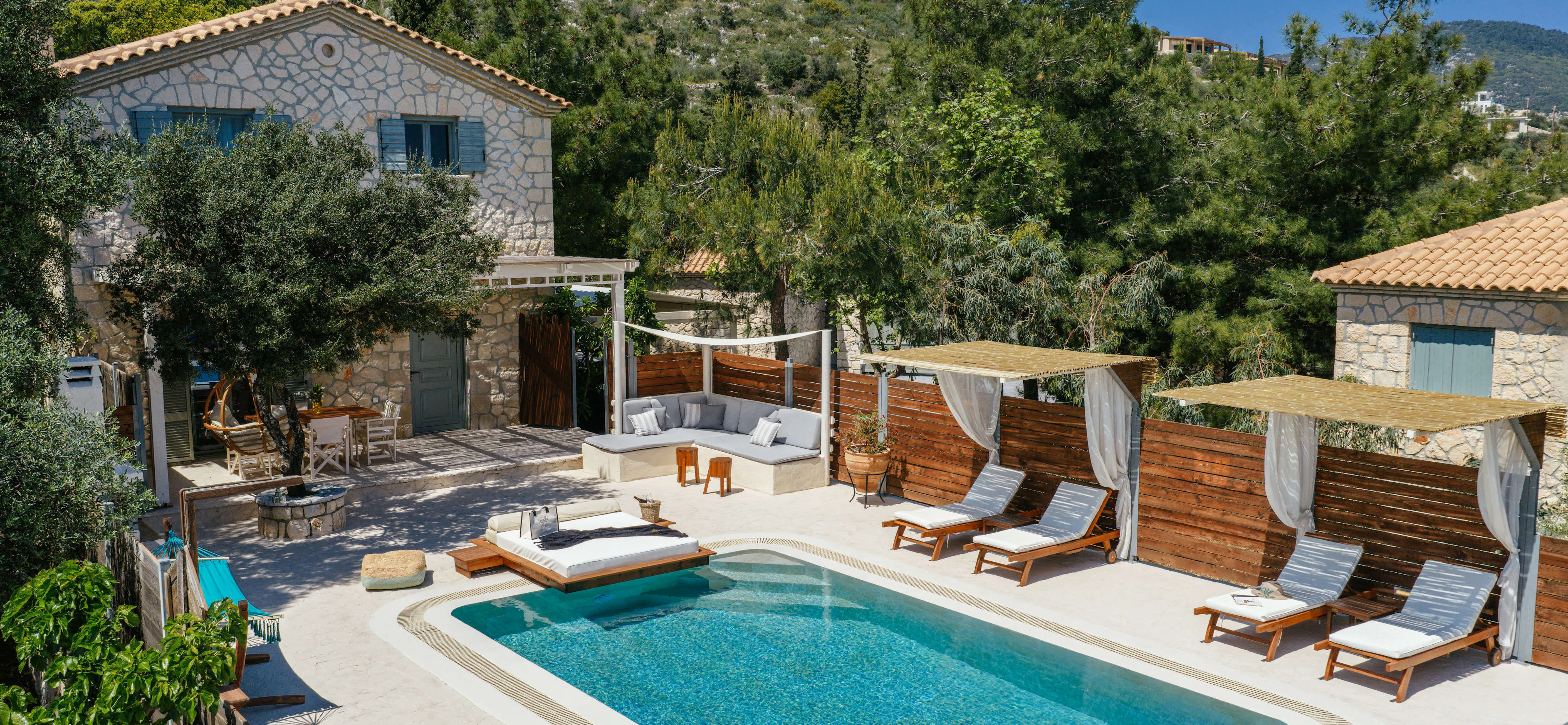 Aerial view of the stone villa and pool terrace, showing shaded outdoor seating, wooden loungers beneath draped cabanas, and a hanging egg chair on the covered deck.