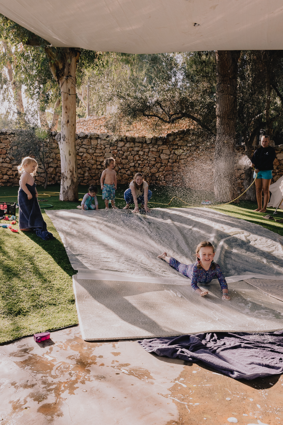 Children play on a soapy slip-and-slide laid across artificial grass in a walled garden, with one girl mid-slide in the foreground and a staff member spraying a hose to keep the mat wet.
