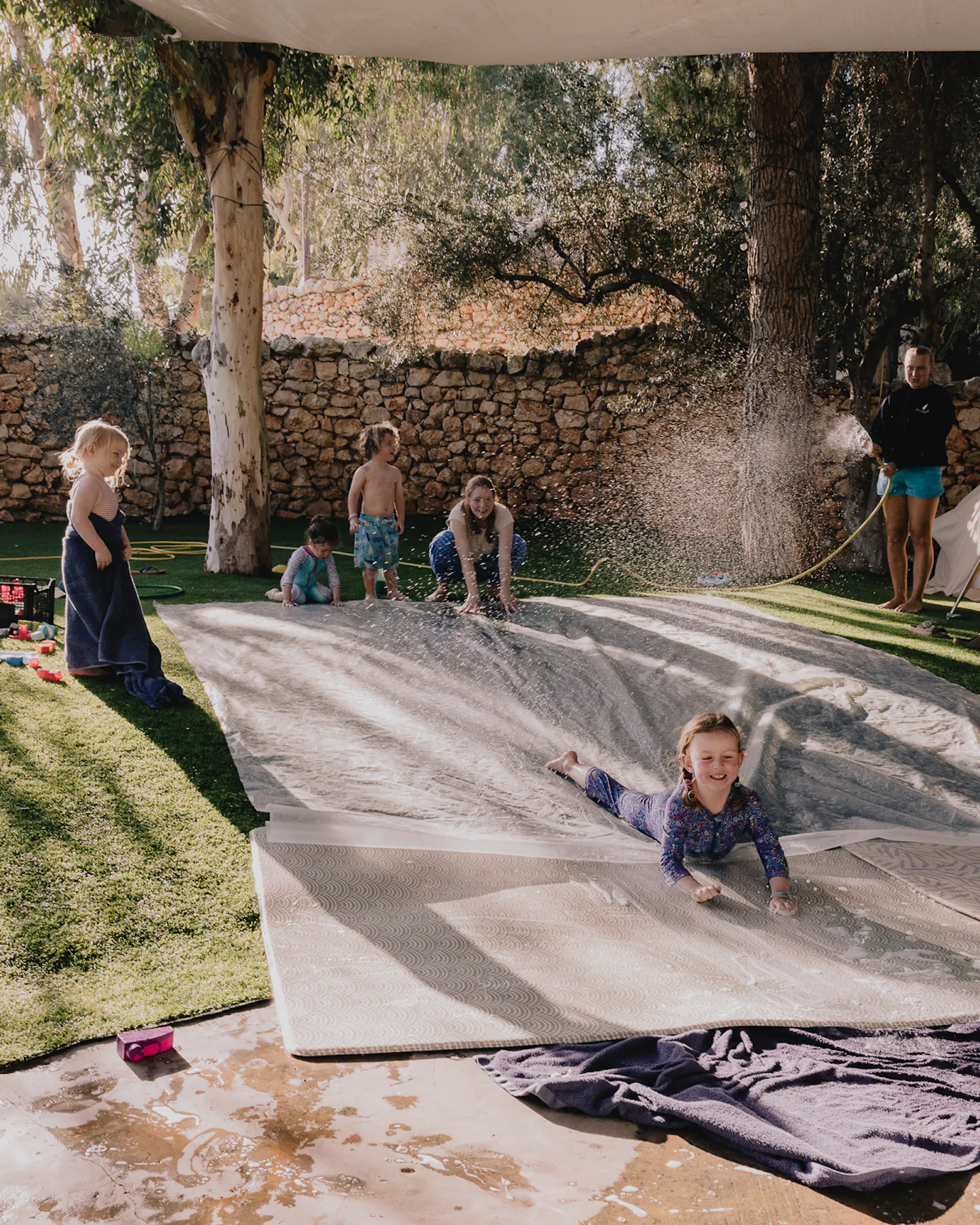 Children play on a soapy slip-and-slide laid across artificial grass in a walled garden, with one girl mid-slide in the foreground and a staff member spraying a hose to keep the mat wet.