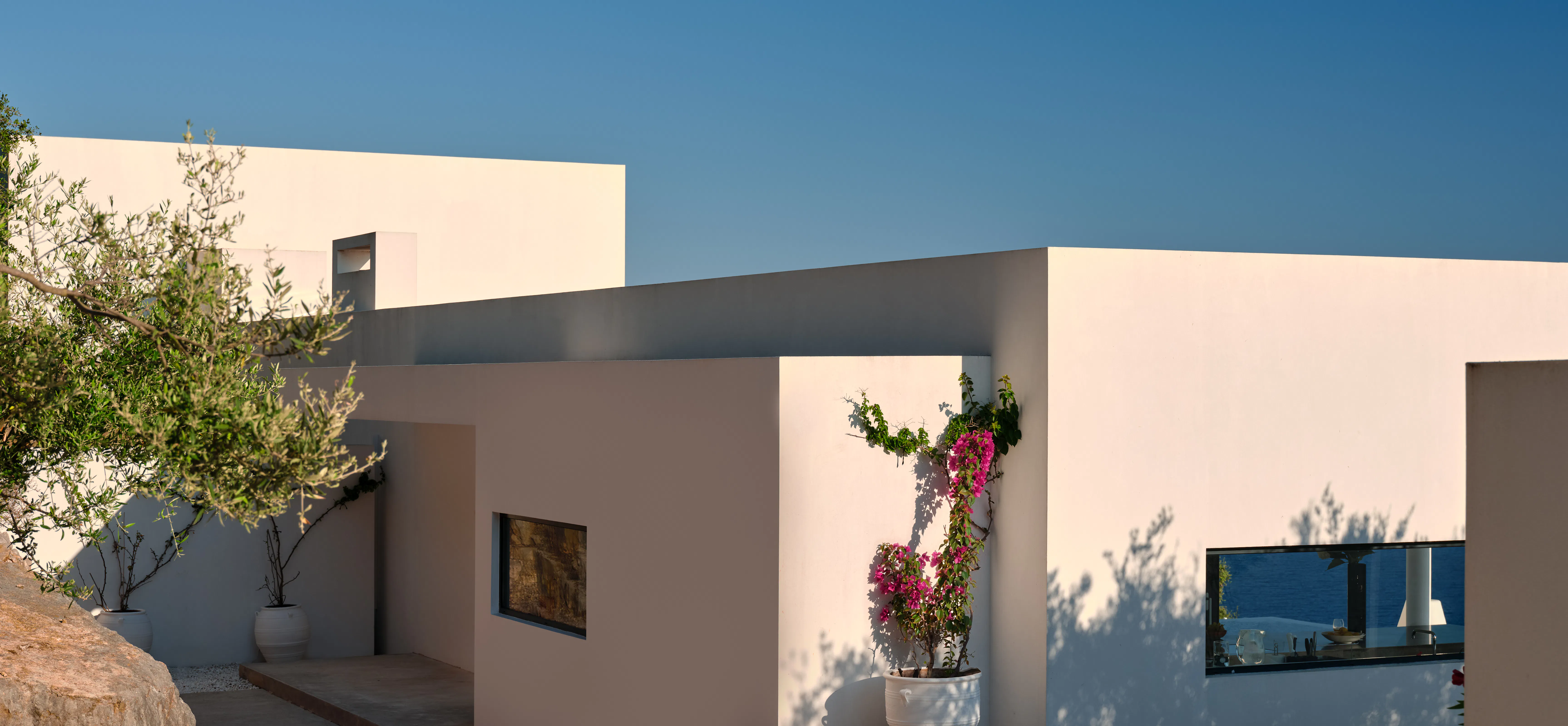 Exterior architectural detail of the villa's white walls and potted bougainvillea, with an olive tree and glimpse of the sea
