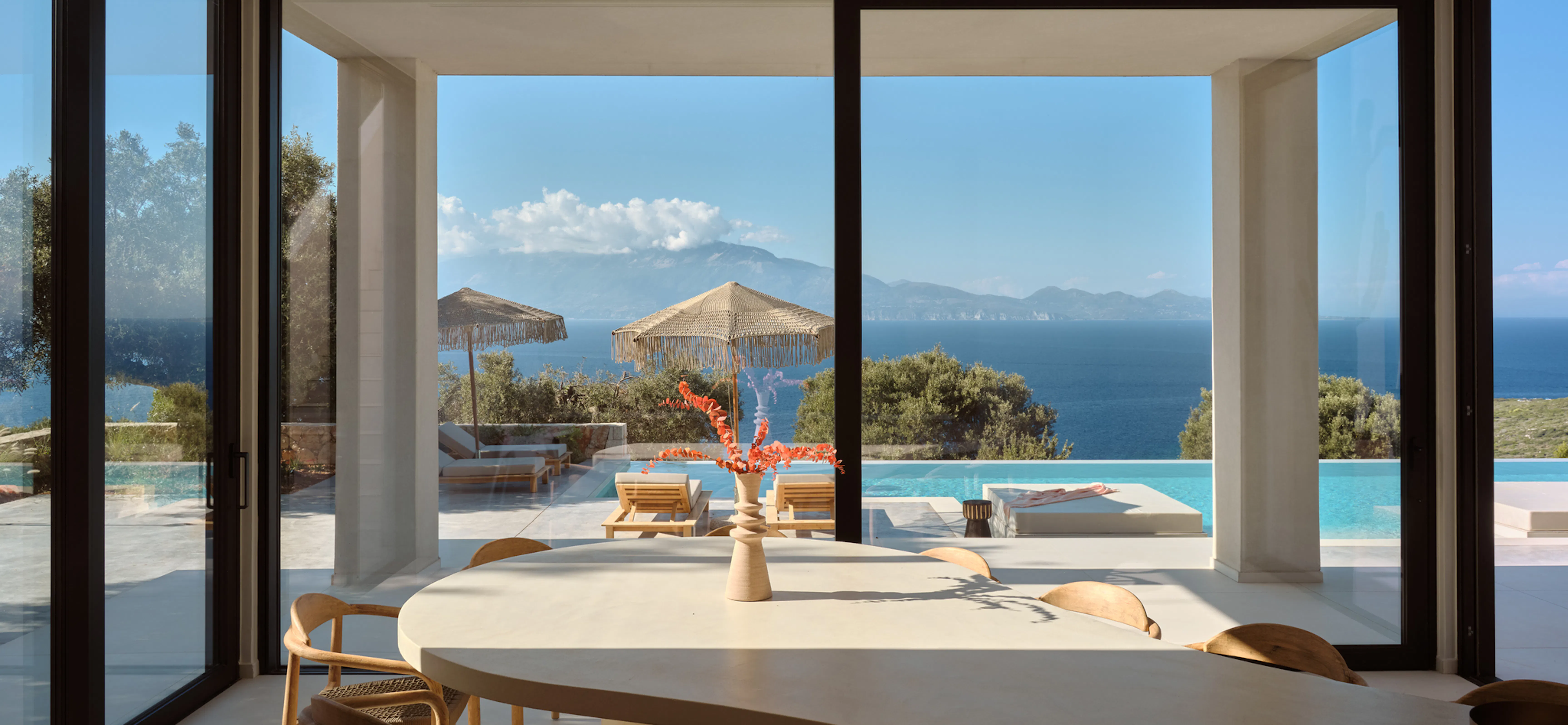 Dining table set beneath a vase of dried branches, glass doors framing the pool terrace, fringed umbrellas, and sea view beyond.