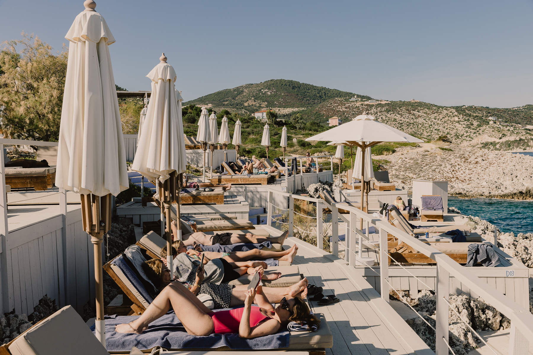 Guests relax on cushioned sun loungers along a whitewashed timber sundeck lined with furled cream umbrellas, with a lush green hillside dotted with villas and a turquoise cove visible beyond the rocky coastline.