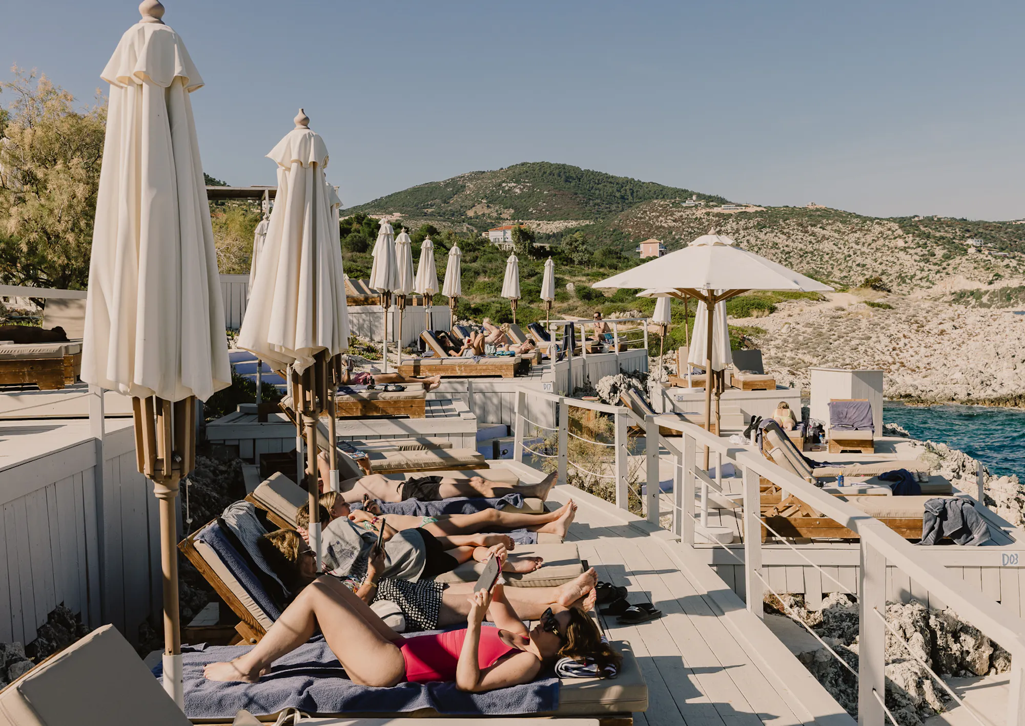Guests relax on cushioned sun loungers along a whitewashed timber sundeck lined with furled cream umbrellas, with a lush green hillside dotted with villas and a turquoise cove visible beyond the rocky coastline.